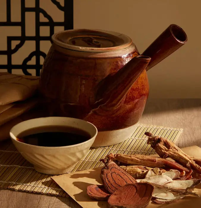 Traditional Chinese herbal medicine with a clay teapot, decoction bowl, and dried herbs on bamboo mat